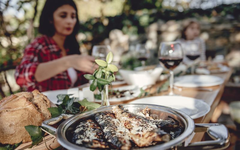 mesa de almoço com um prato de uma receita de peixe em conserva e taças de vinho e uma mulher ao fundo comendo